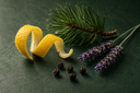 Lemon peel, pine needles, lavender, and peppercorns arranged on a dark green surface.